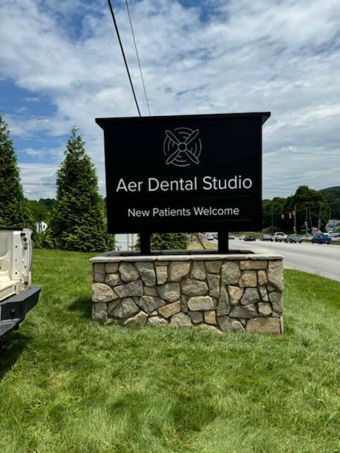 Sign for Aer Dental Studio with black background, logo, and text on a stone base, beside a road. 