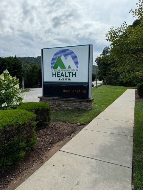 Sign for Mountain Valley Health in Leicester, featuring a blue mountain logo and green text on a white background. The sign is in front of a grassy lawn.