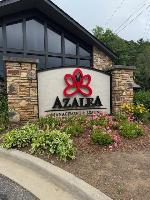 Sign for Azalea Management & Leasing with large red flower logo above the name, surrounded by stone columns and landscaping.