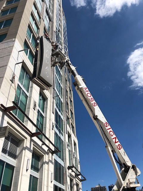 A boom lift elevates a sign on a high-rise building. The lift arm is labeled 