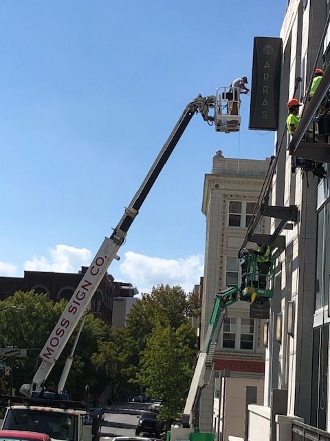 Workers install a sign on a building using a cherry picker and scaffolding. The sky is blue.