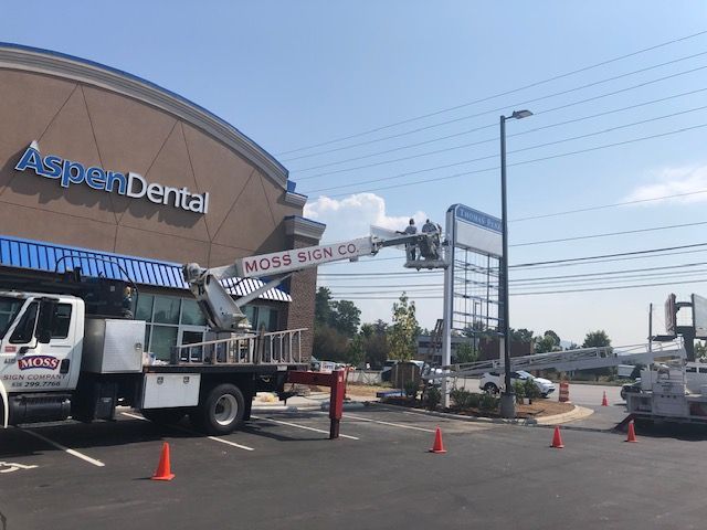 A Moss Sign Co. truck with a lift is installing a sign at Aspen Dental. Orange cones mark the work area in a parking lot.