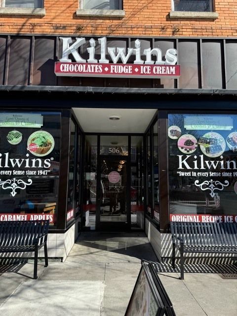Kilwins candy store front with a dark entrance, a brick facade, and signage advertising chocolates, fudge, and ice cream.
