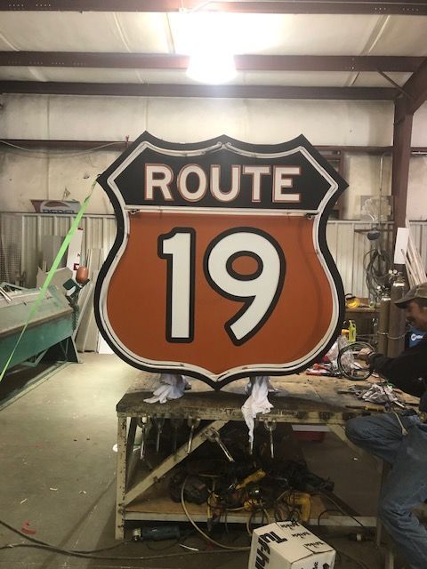 Large Route 19 sign, orange and white, with a black border, sits on a workbench. A person works nearby.