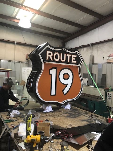 A large Route 19 sign in a workshop, being worked on by a person. The sign is a shield shape, orange and black with white text.