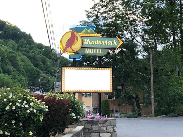 A sign for the Meadowlark Motel with a cardinal illustration and an arrow, with a blank sign below. Lush green hillside in background.
