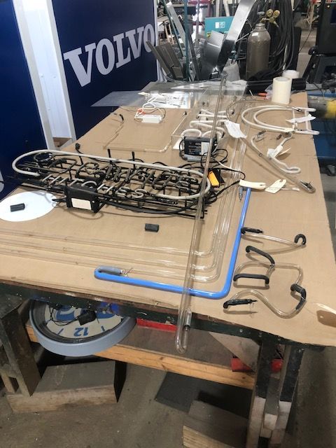 A workbench with various parts of a neon sign, including tubes, wiring, and transformers, alongside a Volvo sign.