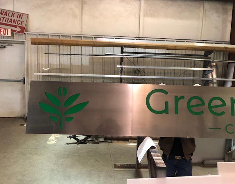A large, brushed metal sign being worked on indoors. It features a green leaf logo and the words 