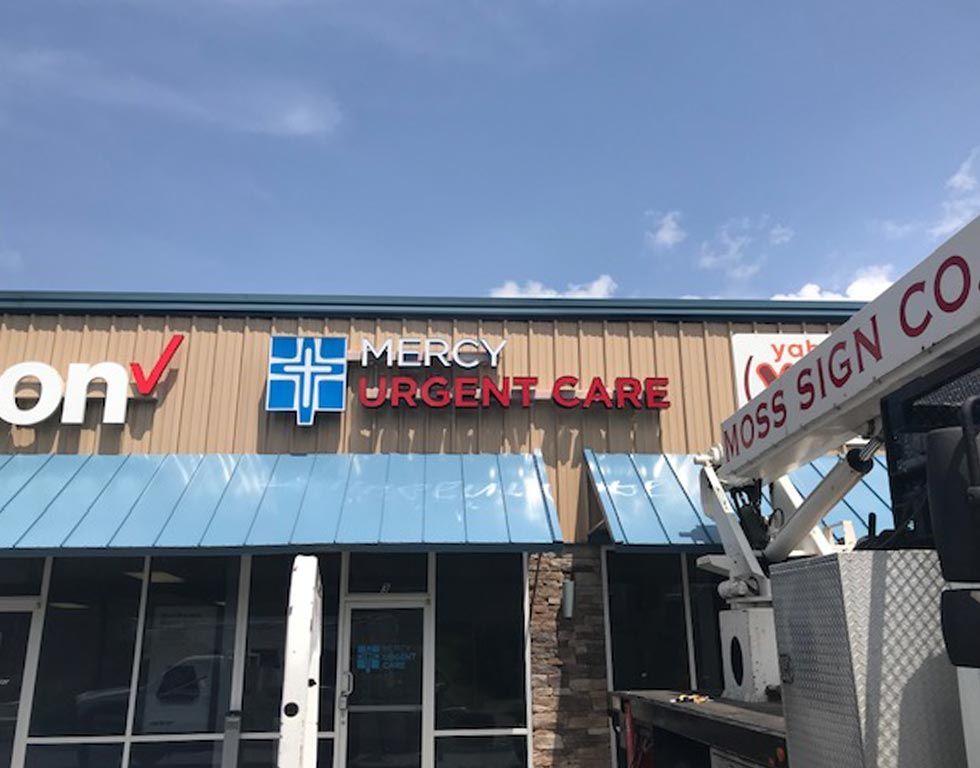A sign for Mercy Urgent Care with a blue cross logo, in red lettering, is being installed at a strip mall under a blue sky.