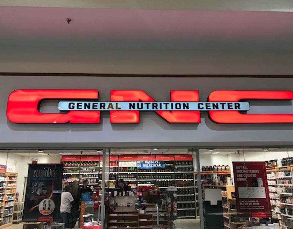 The entrance to a GNC store with a lit-up red sign above the doors, advertising 