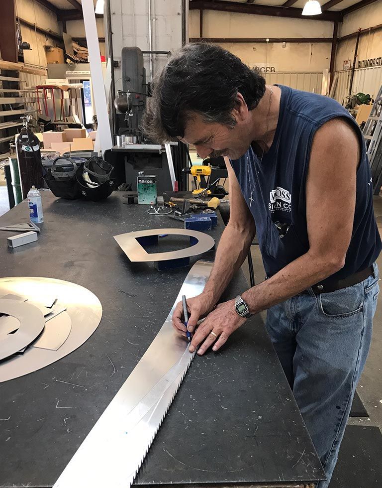 Man in workshop marking metal with a pen. He wears a sleeveless shirt and jeans, working at a metal table with cut metal pieces.