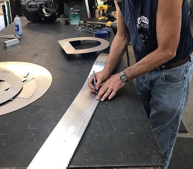 Man in a shop marks a long metal strip with a pen on a metal table, with cut metal shapes nearby.