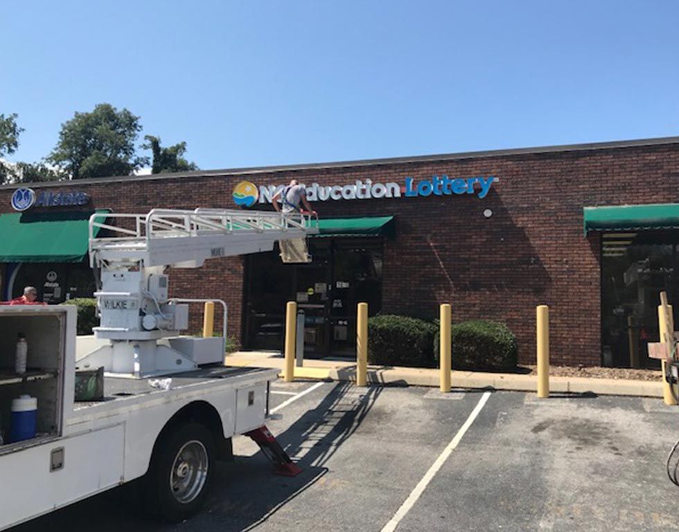 A work truck with a lift platform is servicing the sign for the New Education Lottery on a brick building.