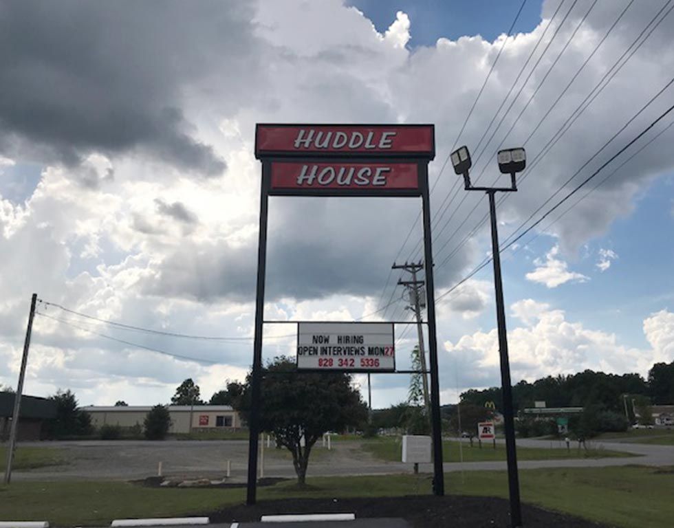 Huddle House sign against a cloudy sky, with hours and a phone number listed below.