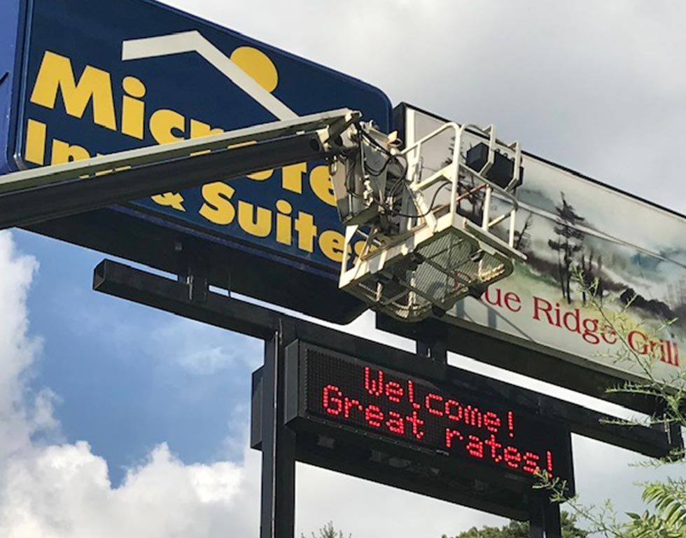 A person in a lift bucket works on a large outdoor sign for a hotel and grill. The sign has a digital display with a welcome message.
