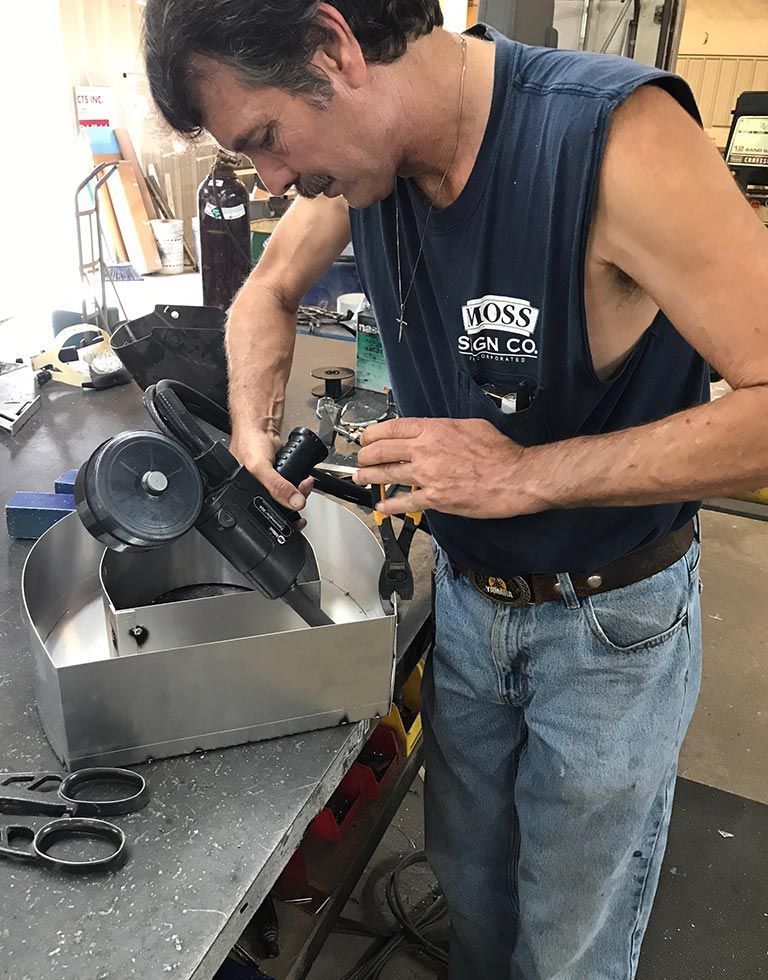 Man welding metal box in a workshop. He wears a blue tank top and jeans while working at a metal table.