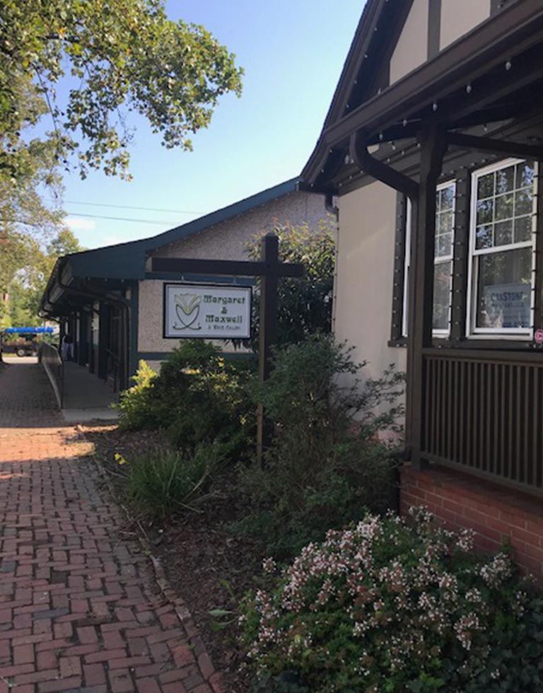 A sign for Maryanne & Friends is in front of a brick building. Lush bushes and a brick pathway lead to the storefront.