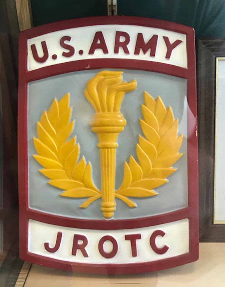 U.S. Army JROTC sign with a gold torch and laurel leaves on a gray background, framed with red and white borders.
