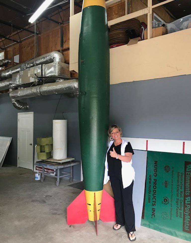Woman giving thumbs up next to a tall, green and yellow rocket in a workshop.