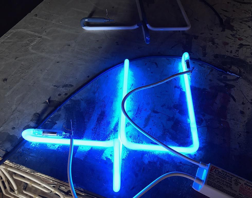 Blue neon light shaped like a plus sign illuminated on a dark surface. Wires and a light ballast are visible.