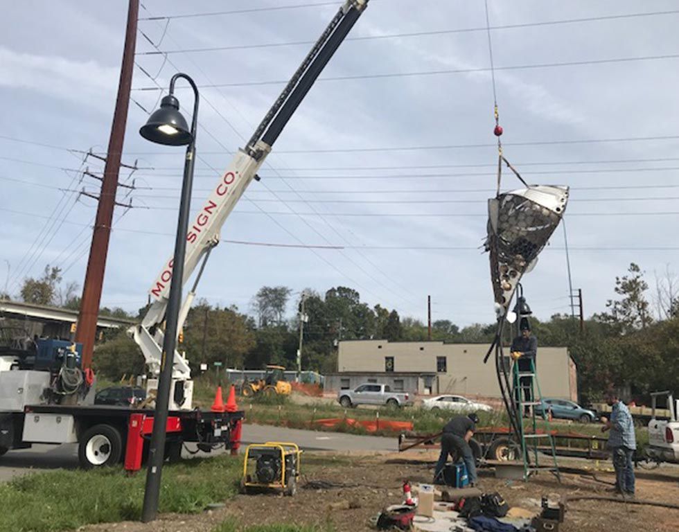 A crane lifts a sign with a curved shape. Workers are on the ground near a power pole in an outdoor setting.
