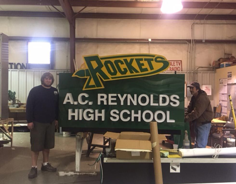 Two men stand by a large, finished green sign for A.C. Reynolds High School, which includes the word 