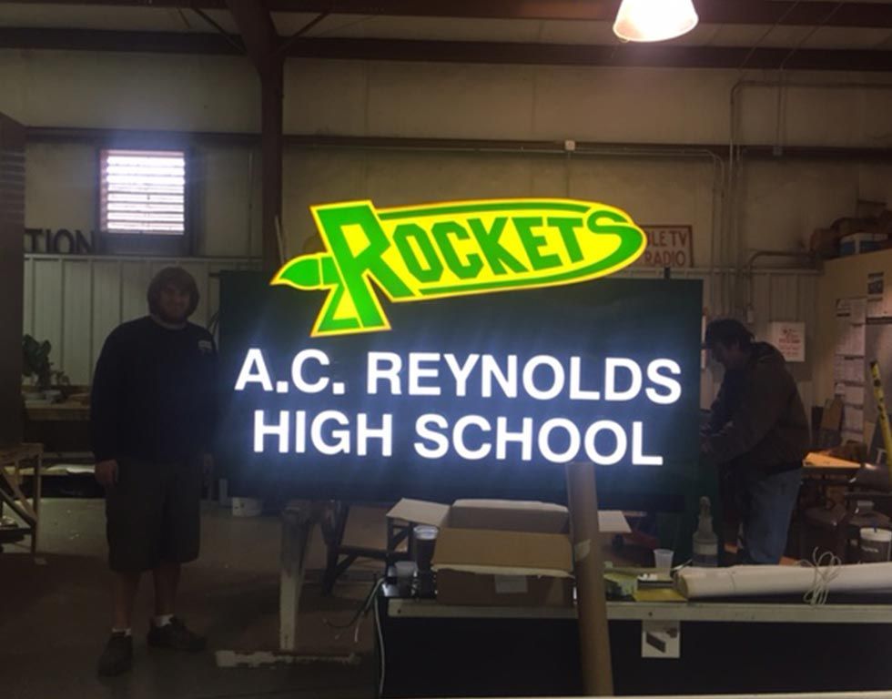 Two men pose with a large LED sign for A.C. Reynolds High School, featuring the school's 