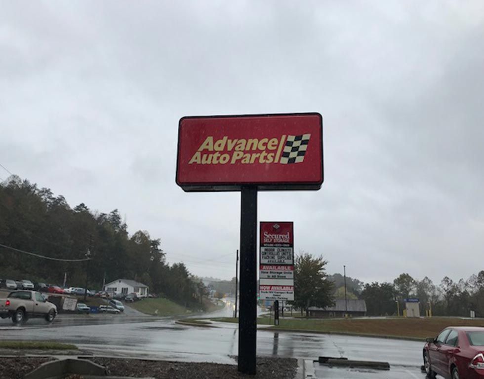 Advance Auto Parts sign in a red box with a checkered flag, standing on a pole in front of a wet road and overcast sky.