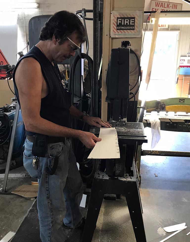 A man in a workshop using a band saw to cut a piece of white wood. He wears glasses and jeans.