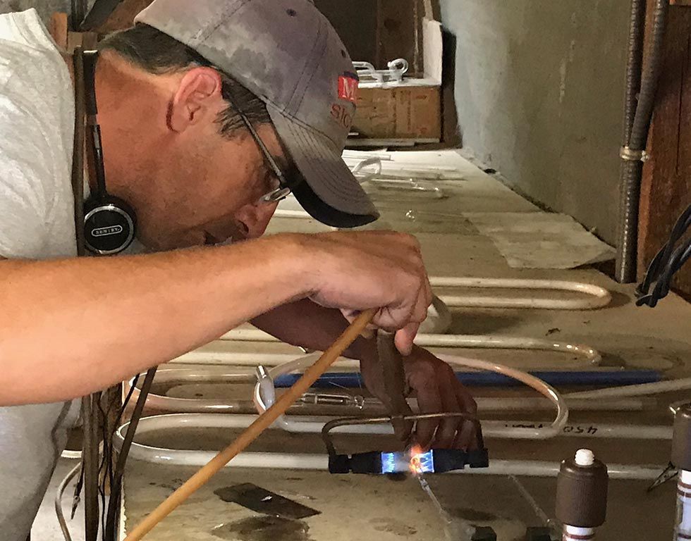A man in a baseball cap soldering pipes in a basement. He's holding a torch, focused, with headphones around his neck.