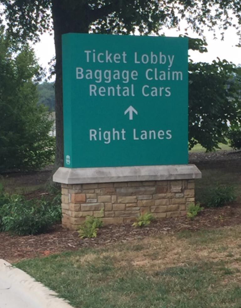 Green signpost with white text pointing towards Ticket Lobby, Baggage Claim, and Rental Cars. Sign is set on a brick and stone base, instructing drivers to use 