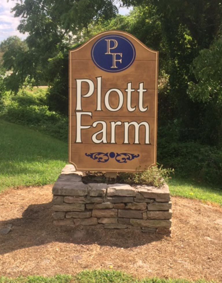 Sign for Plott Farm, with a blue and gold emblem on top, against a background of greenery. The sign is supported by a stone base.