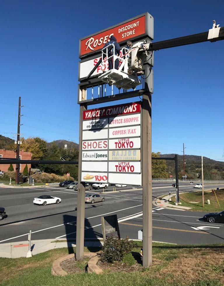 A tall shopping center sign being worked on from a lift, with various store names displayed. Sunny day with a highway and buildings in the background.