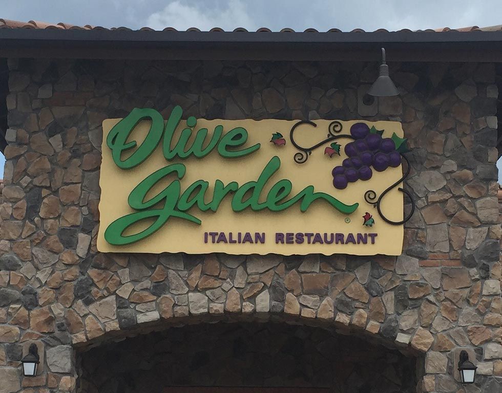 Olive Garden restaurant sign, green cursive lettering on tan background with stone facade.
