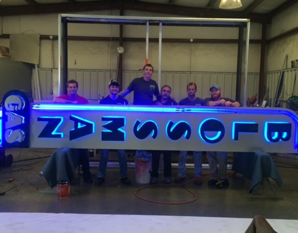 Six people pose with a lit-up, blue neon sign reading 