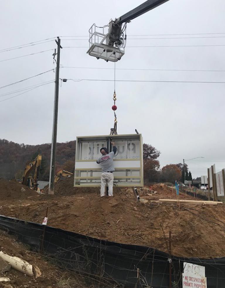 A man stands near a construction site, holding a rectangular panel suspended by a crane. A crane operator is overhead.