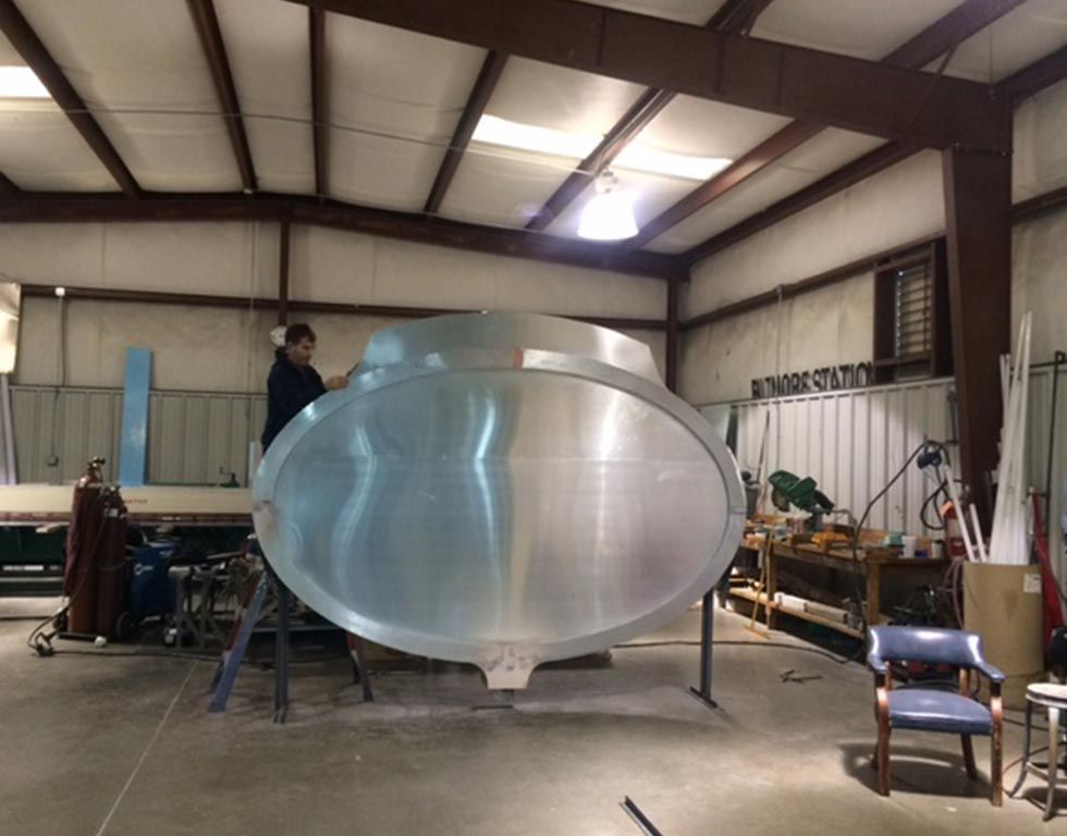 Two people working on a large, oval-shaped metal object in a workshop. The object is on a stand.