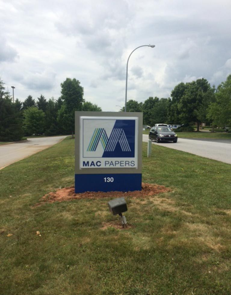 Sign for MAC PAPERS at 133, blue and silver logo on a gray square, roadside grass, cloudy sky, and a car.