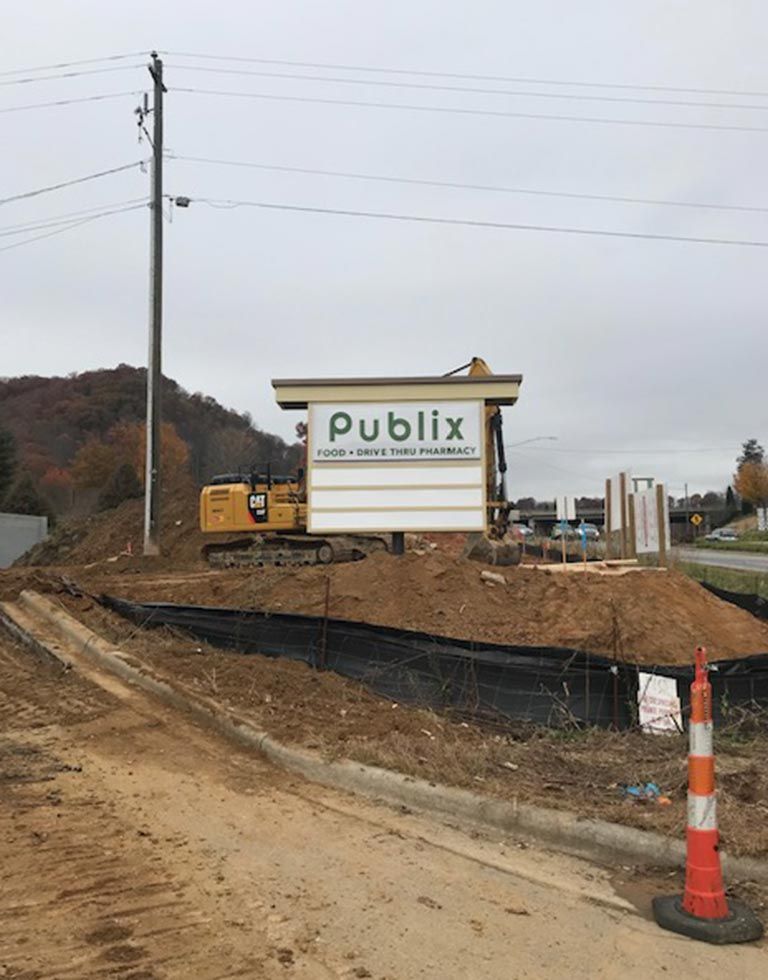 A construction site with a Publix sign. An excavator works near the sign; surrounding the site are dirt piles and a road.