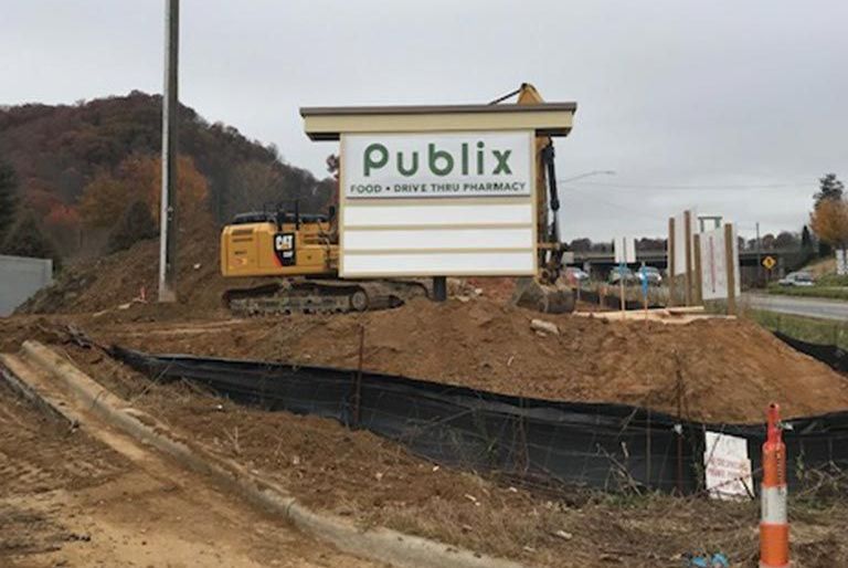 A construction site with a new Publix sign; an excavator sits nearby, with dirt surrounding the sign.