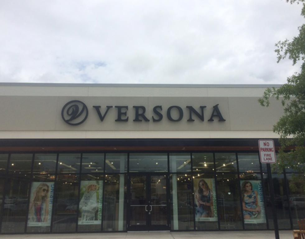 A Versona clothing store with a large sign under a cloudy sky. The front has large windows with mannequins inside.