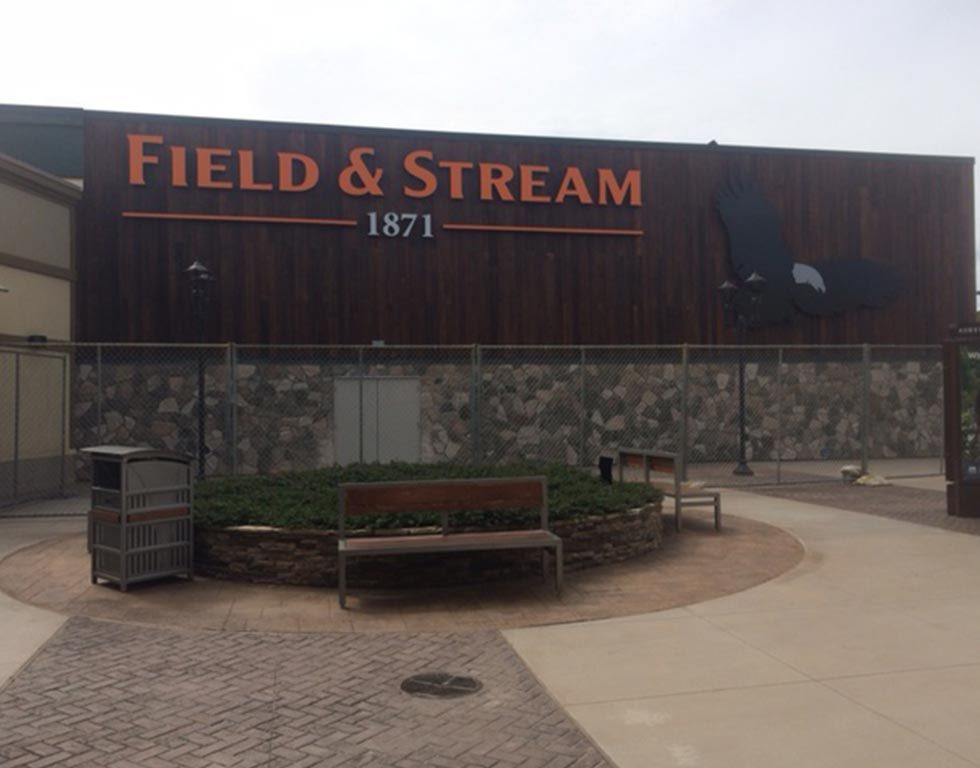 Field & Stream store exterior with a brown facade, orange logo, and a stone wall. Benches and greenery are in front.