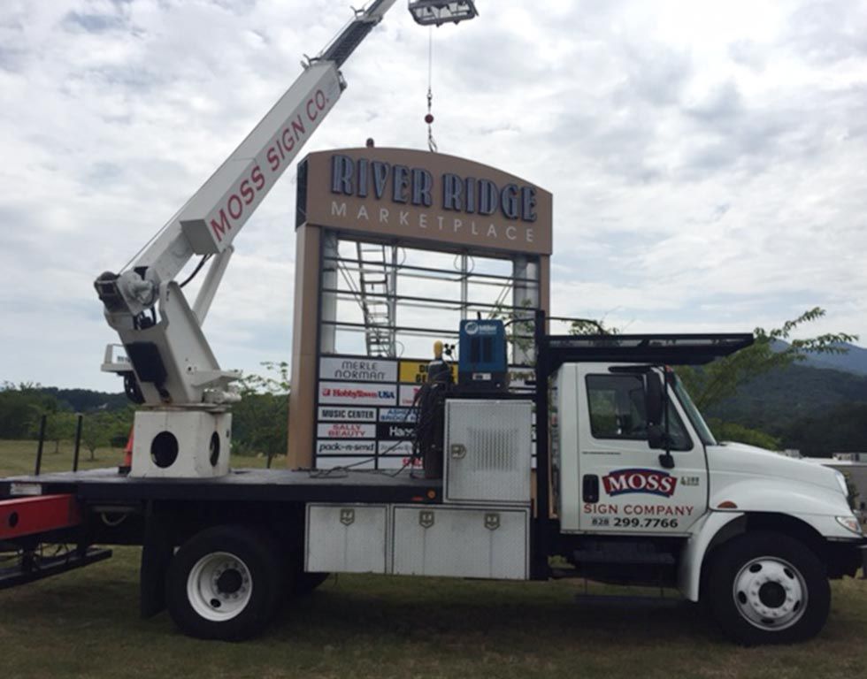 A crane truck from Moss Sign Co. installs a sign for River Ridge Marketplace. The white truck is parked outdoors.
