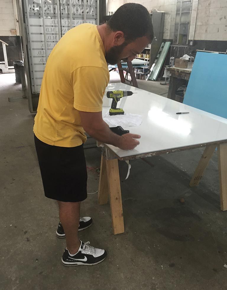 A man in a yellow shirt uses a power drill on a white surface set on sawhorses in a workshop.