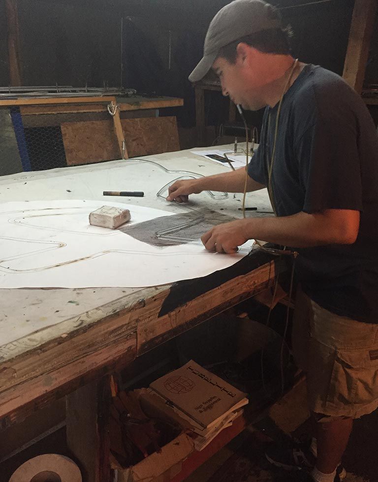 A man in a cap uses a ruler to trace a pattern on a large white paper on a workbench. He’s in a workshop setting.