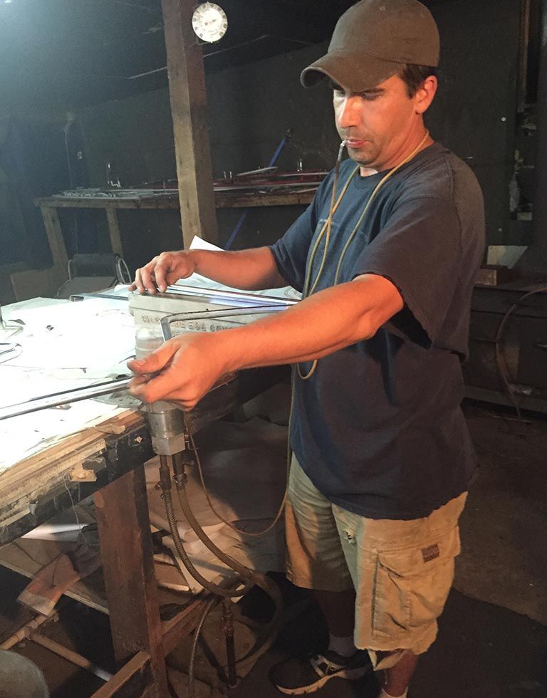 Man in a cap and khaki shorts working at a workbench, possibly a workshop. He’s holding a small item.