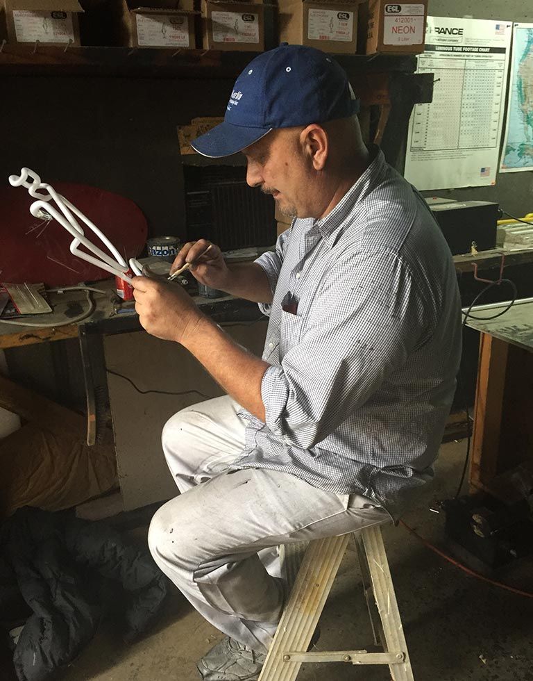 Man in a blue cap and striped shirt seated, working on white metal object. He’s in a workshop with various tools and supplies.