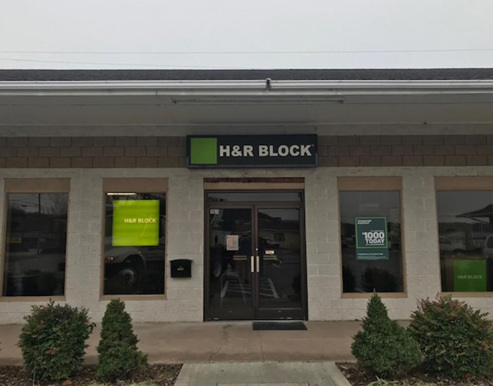 H&R Block storefront with a green and black sign, windows, and bushes. The sky is overcast.