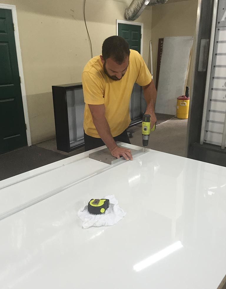 A man in a yellow shirt uses a drill on a white surface in a workshop. A measuring tape sits nearby.