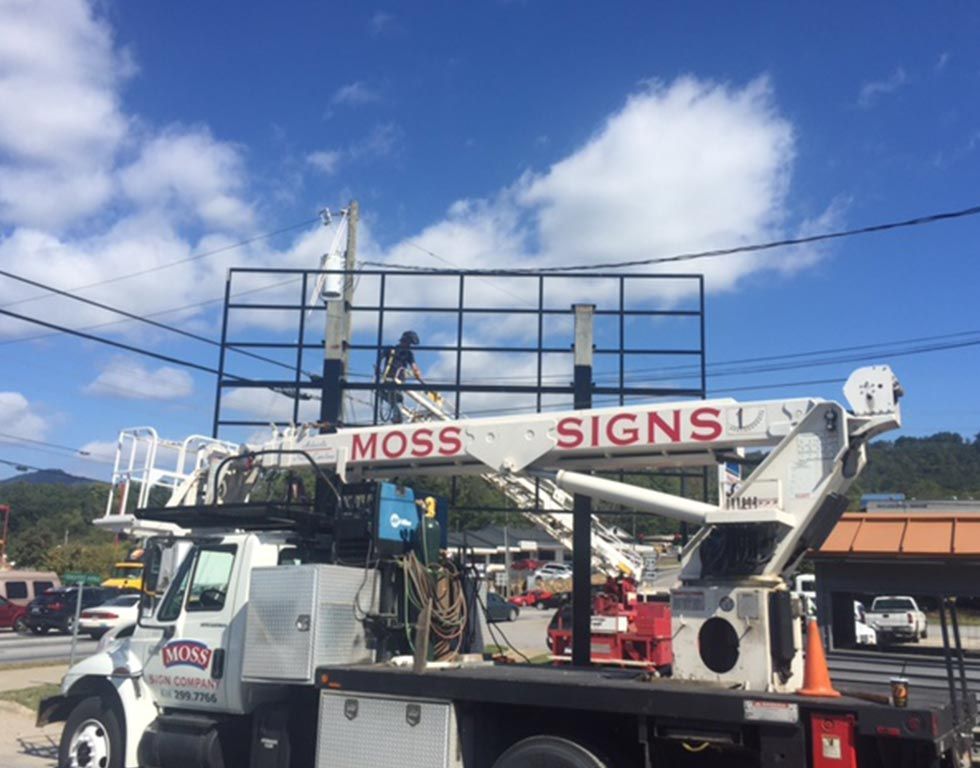 A sign company truck with a raised platform, supporting a person working on a large billboard frame against a cloudy blue sky.